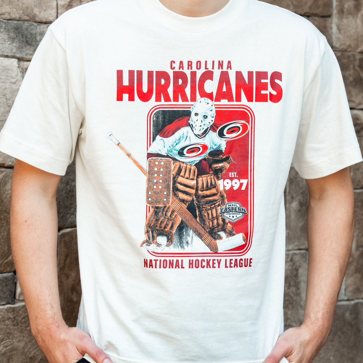Front: White tee, "CAROLINA HURRICANES NATIONAL HOCKEY LEAGUE" in red around rectangular artwork of vintage goalie